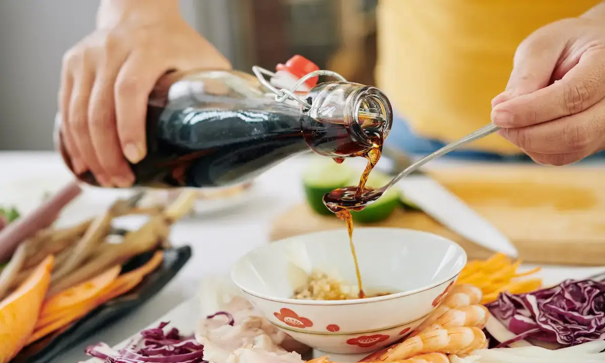 Teriyaki sauce poured from glass bottle onto spoon over bowl with shrimp and vegetables