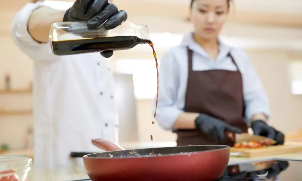 Chef pouring teriyaki sauce into a skillet while assistant chops vegetables