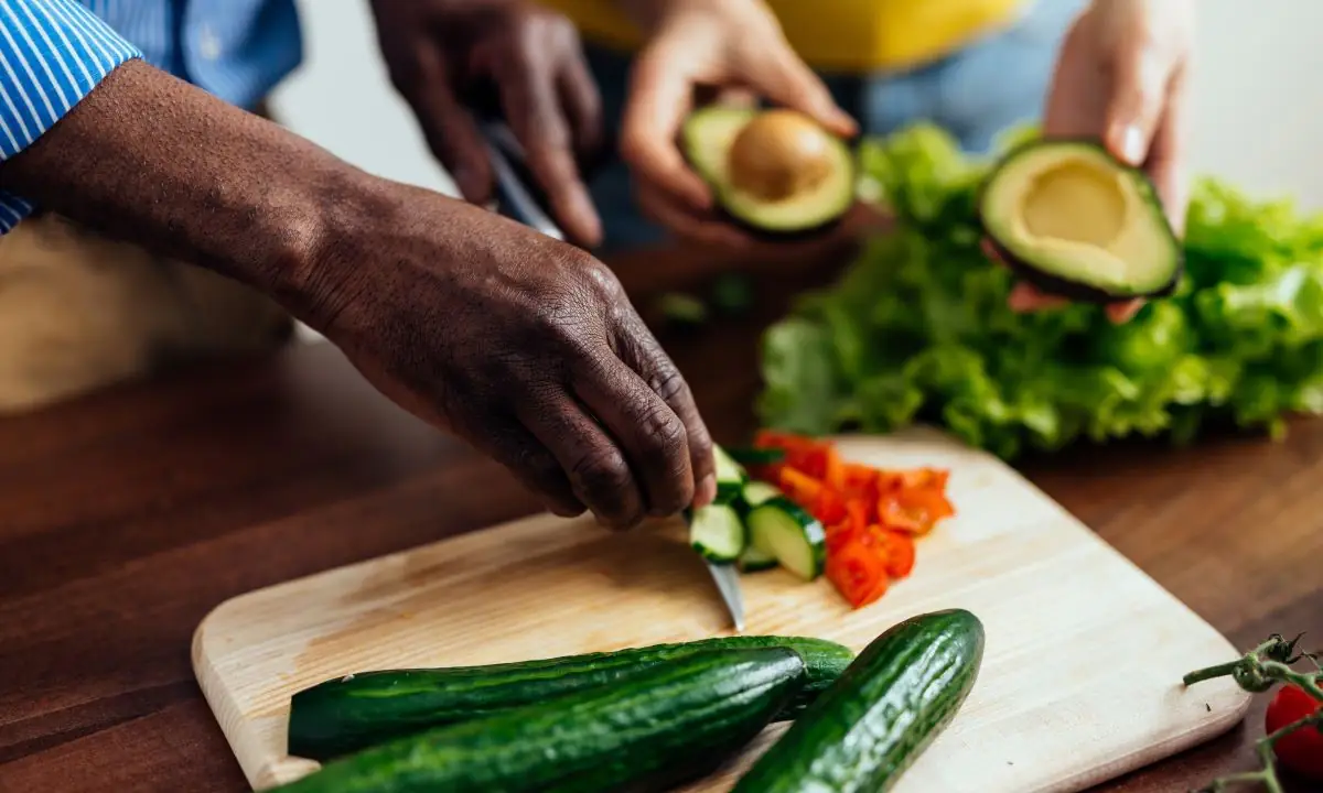 Hands slicing cucumbers with halved avocados and lettuce for teriyaki salmon bowl