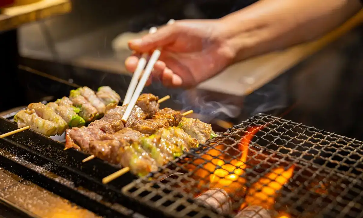 Teriyaki beef skewers grilling over charcoal, hand turning with chopsticks